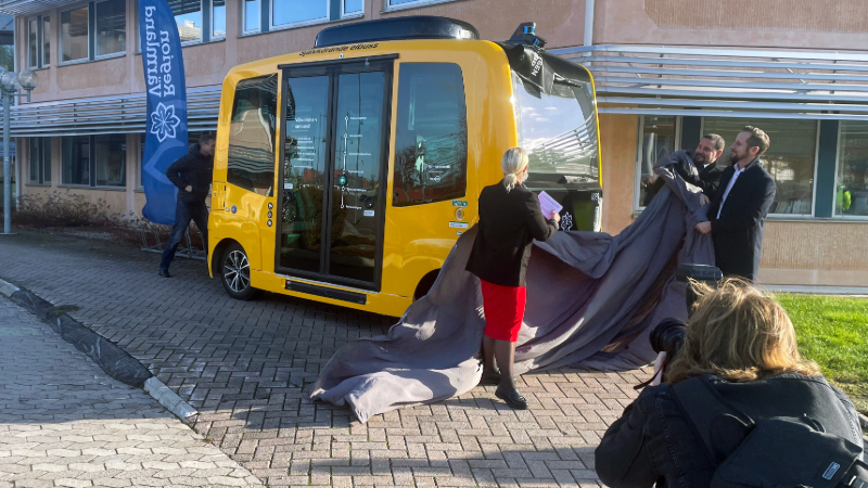 tre personer tar av ett skynke som täckt en liten gul självkörande buss. I bakgrunden syns Regionens hus.