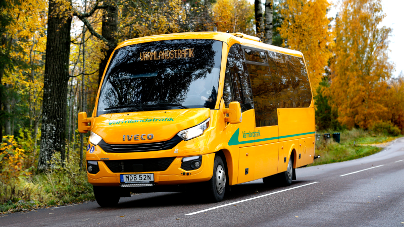 En servicetrafiksbuss som åker på en väg, i bakgrunden ser man träd med höstlöv.