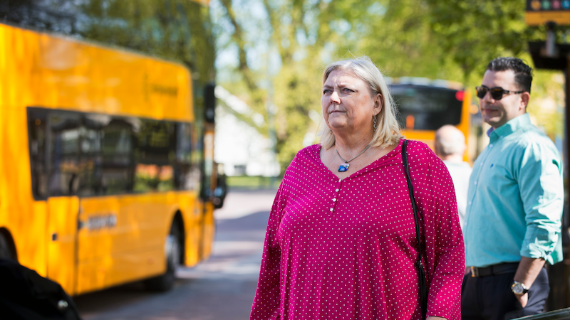 En kvinna som står och väntar på bussen i bakgrunden står en man och en gul buss åker förbi. 